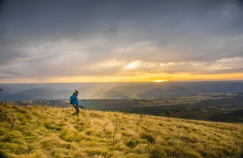 Wanderer mit Rucksack und Trekkingstöcken auf grasigem Kamm bei Sonnenuntergang, weites Tal und goldenes Licht