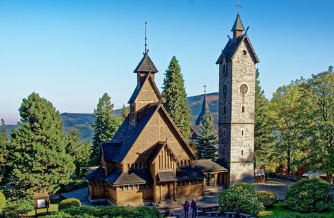 Drewniany kościół typu stavkirke z ostrymi szindelowymi dachami i kamienną wieżą dzwonniczą, wśród sosen i gór; dwoje gości przy wejściu