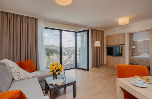 Spacious suite living area with sofa, orange cushions, coffee table with yellow tulips and open book, balcony with deck chairs overlooking forest