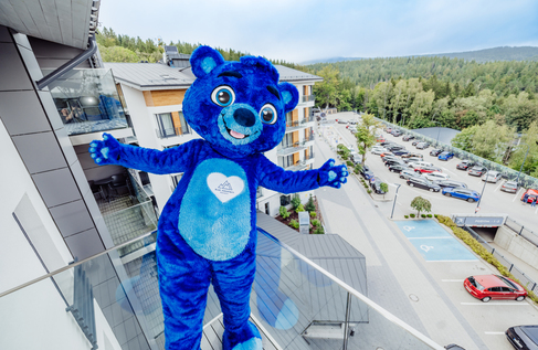 Blue bear mascot on hotel balcony welcoming guests, resort logo on chest, forested mountain view and parking below