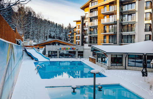 Outdoor heated pool and water slide beside a modern alpine hotel with glass balconies, snow and forest backdrop.