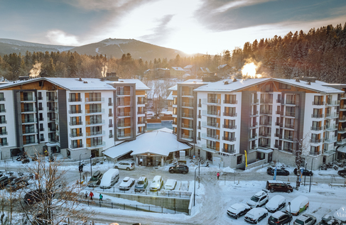 Modern alpine hotel complex with balconies, snowy parking and steam from chimneys, sunrise over forested mountains and distant ski slopes