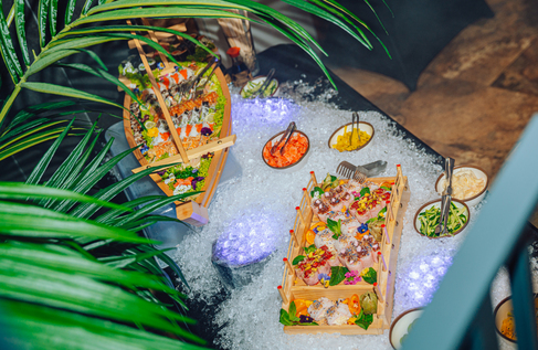 Loďky se sushi a sashimi na drceném ledu, barevné ozdoby, misky s nakládanou zeleninou a zázvorem, tropické listy.