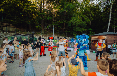 Resort outdoor kids' show with costumed characters and host leading children dancing on a forested evening stage