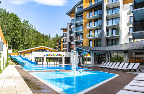 Hotel outdoor pool and aquapark with blue waterslide, central fountain jets, rows of sunbeds and modern wood-clad rooms with balconies facing forest.