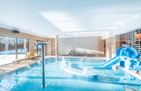 Sunlit indoor hotel pool with water jets, loungers, mushroom fountain and a blue children's elephant slide.