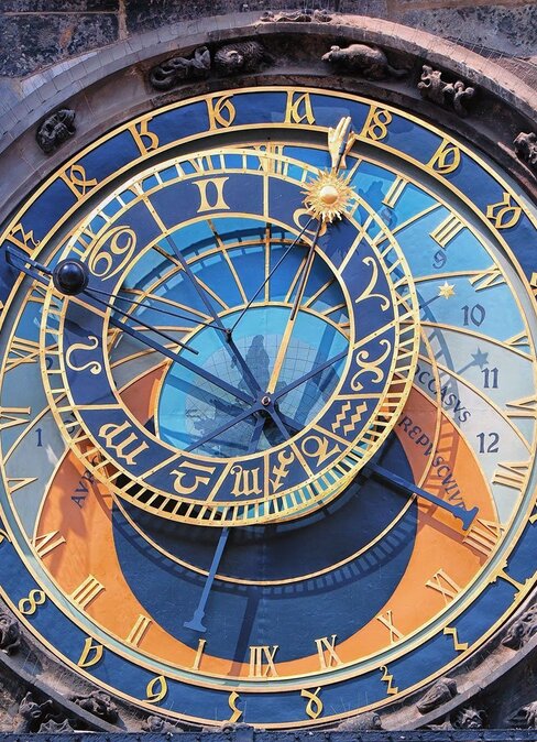 Bogato zdobiony zegar astronomiczny na kamiennej wieży, niebiesko‑złota tarcza z znakami zodiaku, złocone wskazówki i rzeźbione figury, w tym szkielet