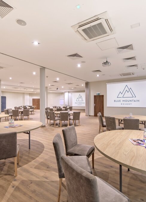 Prostorná konferenční místnost s kulatými stoly, čalouněnými židlemi, projekční plochou s logem Blue Mountain Resort a dřevěnou podlahou.
