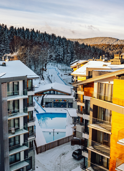 Modern alpine hotel buildings with wood facades, snow-covered roofs and balconies; outdoor pool and aquapark entrance, dense pine forest on the hills