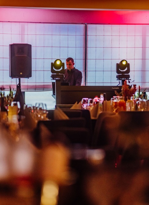 Hotelová lounge s DJ pultem, reproduktory a teplým růžovým osvětlením, prostřené stoly se sklenicemi k večeři.