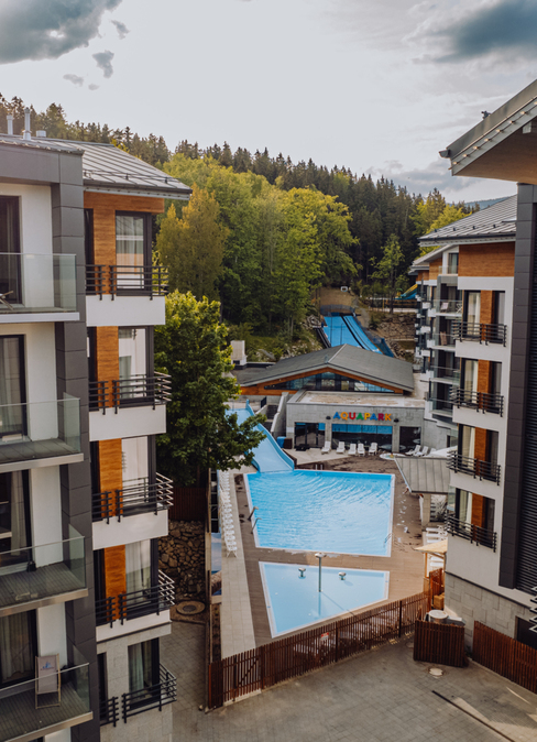 Modern mountain hotel complex with glass balconies, outdoor pool and aquapark slides set against a forested hillside.