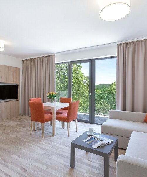 Jasny nowoczesny apartament z salonem: beżowa sofa z pomarańczowymi poduszkami, stół dla 4, TV, balkon z widokiem na las, sypialnia widoczna przez drzwi