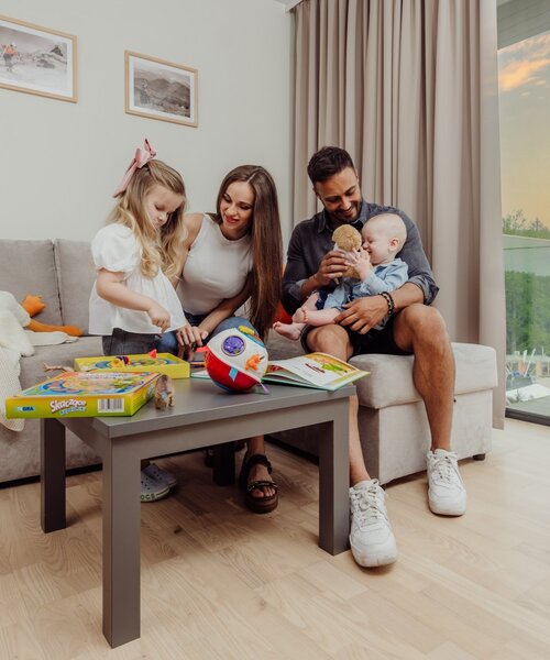 Salon rodzinnego apartamentu, rodzice bawią się z dziećmi i zabawkami na kanapie, przeszklony balkon z widokiem na las o zachodzie słońca, pies