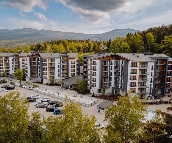 Moderní horské středisko s vícepatrovými budovami a balkony, parkovištěm, dětským hřištěm a výhledem na lesy a hřebeny.