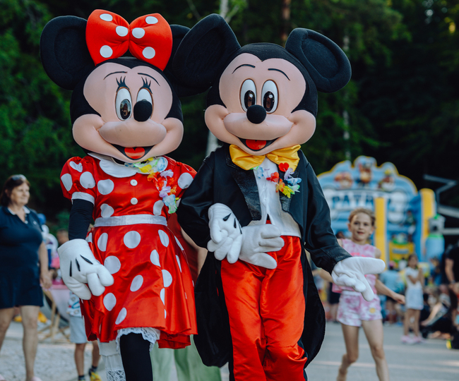 Kostümierte Maskottchen Minnie und Mickey laufen Händchen haltend bei einer lebhaften Familienveranstaltung im Resort.