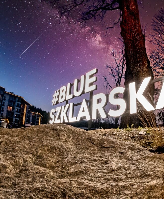 Podświetlony napis #BLUE SZKLARSKA na skalnym pierwszym planie, przytulny górski hotel z ciepłymi lampkami pod gwiaździstym niebem