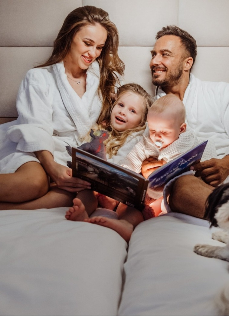 Family in white robes on a plush king bed with padded headboard, reading bedtime story by soft light; pet-friendly suite