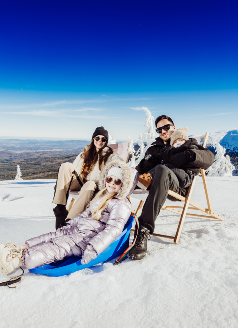 Family relaxing on deck chairs with a child in a sled on a sunny snowy alpine summit and panoramic mountain views