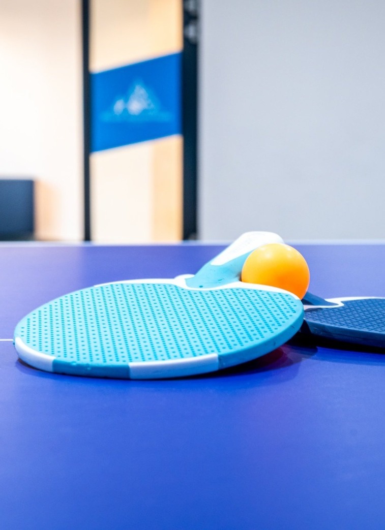 Dvě modré pálky na stolní tenis a oranžový míček na modrém stole v světlé hotelové herně.