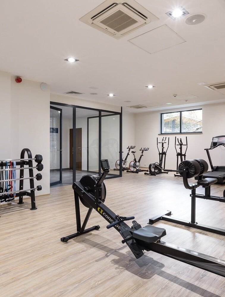 Světlé hotelové fitness s veslovacím trenažérem, běžeckými pásy, rotopedy, činkami a světlou dřevěnou podlahou