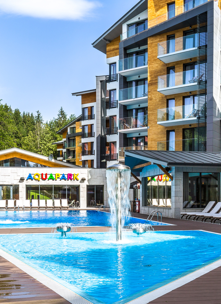 Hotel outdoor pool and aquapark with blue waterslide, central fountain jets, rows of sunbeds and modern wood-clad rooms with balconies facing forest.