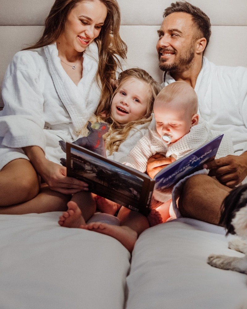 Rodzina w hotelowym, przytulnym łóżku w szlafrokach czyta wieczorną bajkę dzieciom, mały pies odpoczywa.