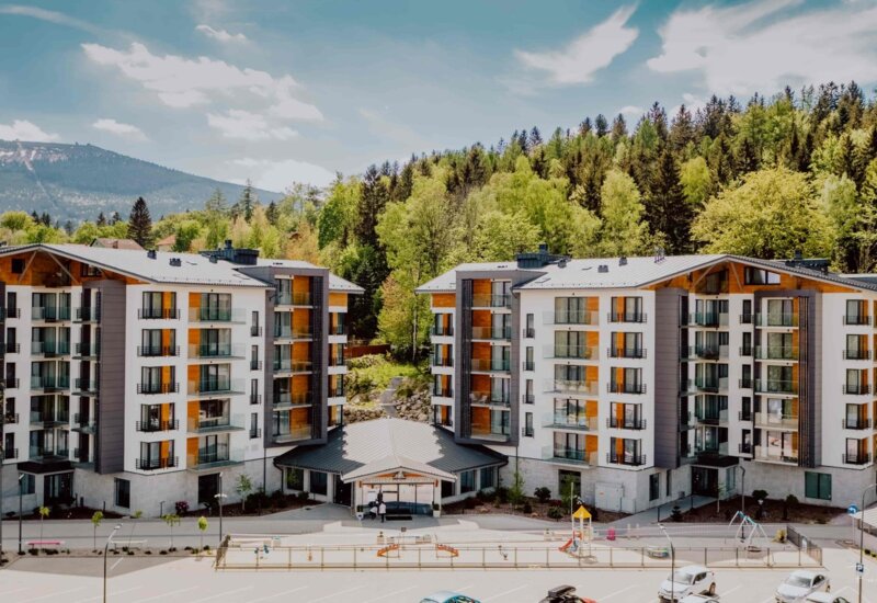 Moderní horské letovisko s dřevěnými prvky, skleněnými balkony, hlavním vchodem, dětským hřištěm, parkovištěm a zalesněnými svahy v pozadí.