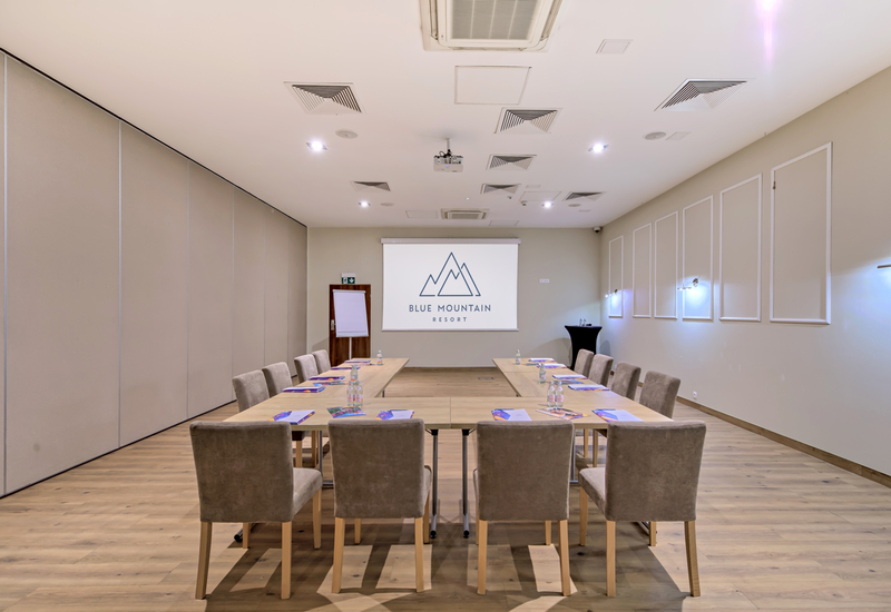 Sala konferencyjna w układzie U, tapicerowane krzesła, ekran z logo 'Blue Mountain Resort', flipchart i woda.