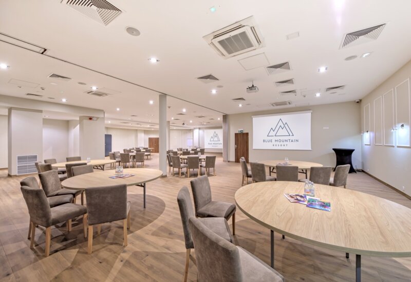 Przestronna sala konferencyjna z okrągłymi stołami, tapicerowanymi krzesłami, ekranem z logo Blue Mountain Resort i drewnianą podłogą.