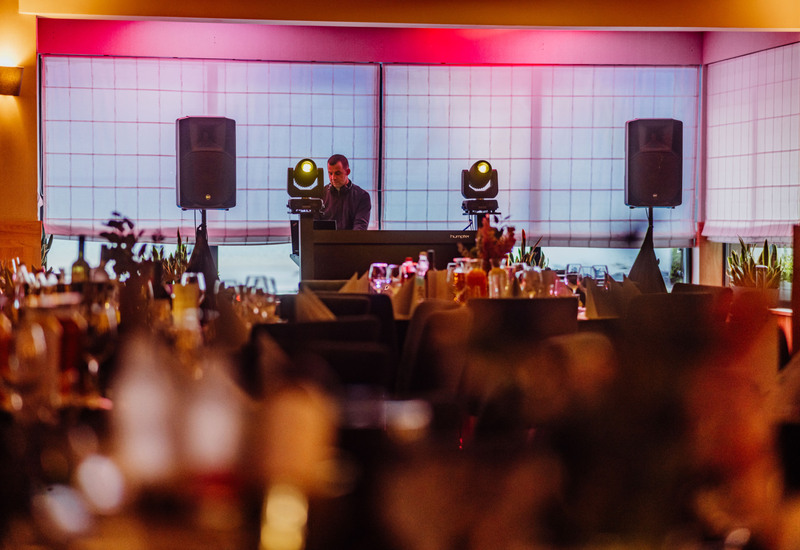 Hotelowa sala z DJ-em, głośnikami i różowym oświetleniem, nakryte stoły z kieliszkami gotowe na kolację.