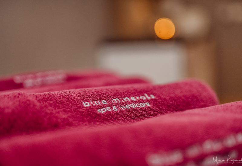 Miękkie fuksjowe ręczniki spa z wyszywanym napisem „blue minerals spa & medicore”, miękka faktura i rozmyte światło świecy tworzą relaksujący klimat