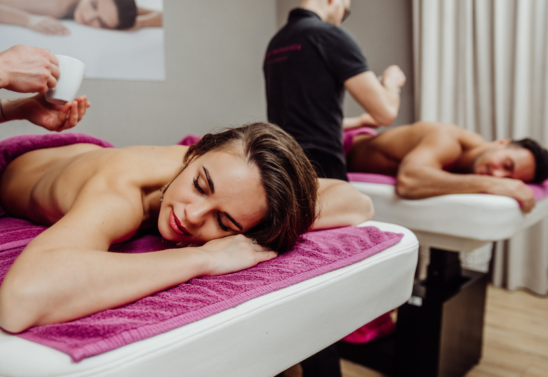 Masaż dla par w hotelowym spa na sąsiednich leżankach z fioletowymi ręcznikami, olejek aplikowany, relaksująca atmosfera.