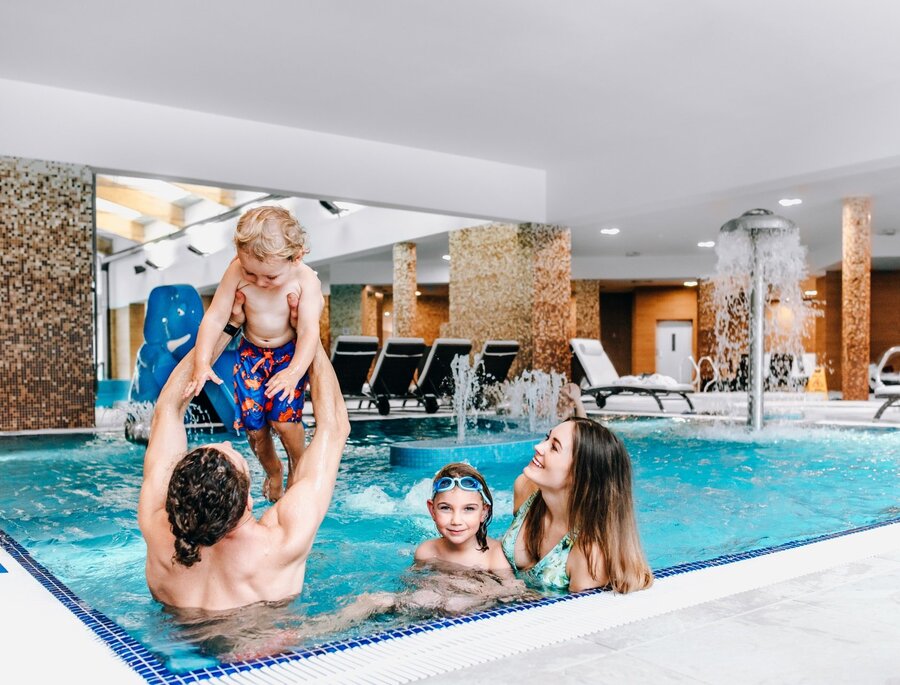 Familie spielt im hoteleigenen Hallenbad – Vater hebt Kleinkind, Mutter und Kind lächeln am Springbrunnen und Liegen.