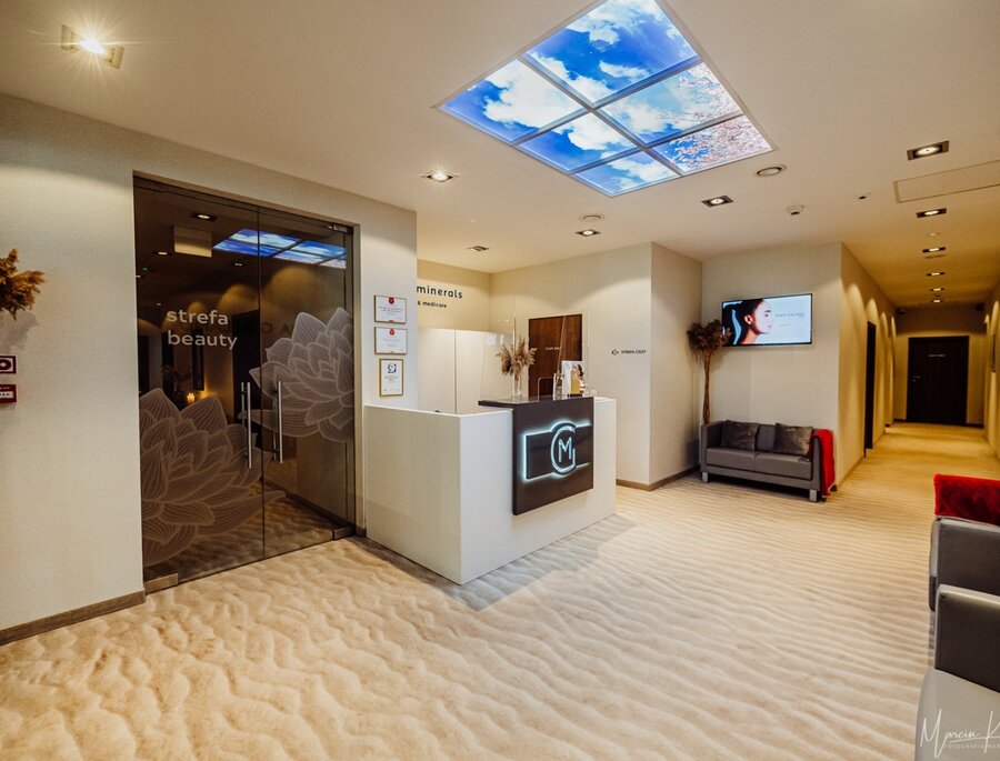 Spa-Empfang mit weißer Theke, Glastür mit Aufschrift 'strefa beauty', himmelsbeleuchteten Deckenpanelen und bequemen Sofas.