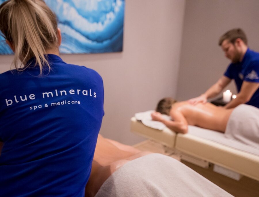 Równoległe masaże w przytulnym, przyciemnionym pokoju spa, terapeuci w niebieskich koszulkach 'Blue Minerals', świeczki i obraz