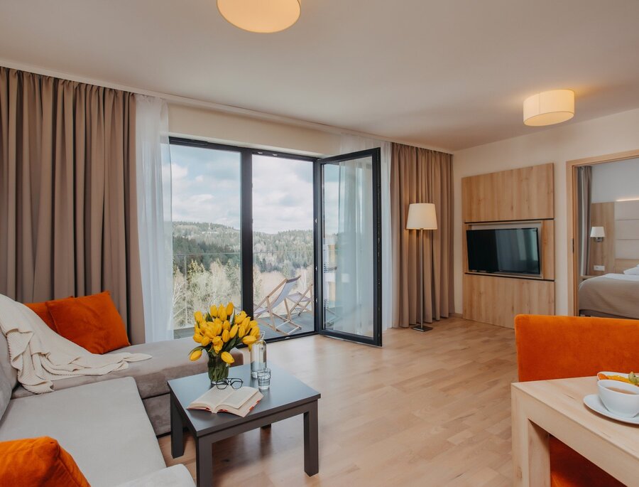 Prostorná suita s obývacím koutem, pohovkou s oranžovými polštáři, stolkem se žlutými tulipány a otevřenou knihou, balkon s lehátky a výhledem do lesa