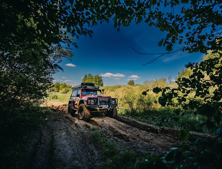 Verschlammter 4x4-Geländewagen auf zerfurchtem Waldweg, eingerahmt von Laubdach und einer sonnigen Lichtung