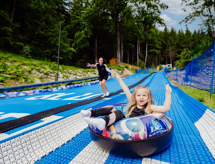 Kind lacht in einem Luftreifen auf blauem Sommer-Tubing-Kanal im bewaldeten Bergresort, Betreuer rennt dahinter