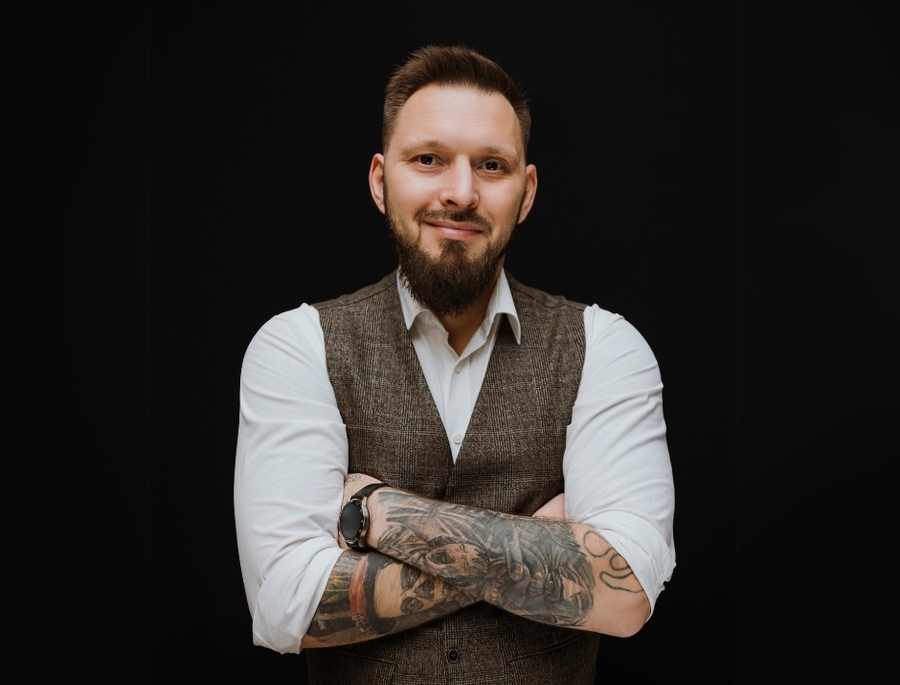 Zuvorkommender Hotelmitarbeiter mit Bart und Ärmel‑Tattoos, in Tweedweste und aufgekrempeltem weißen Hemd, verschränkte Arme, freundliches Lächeln