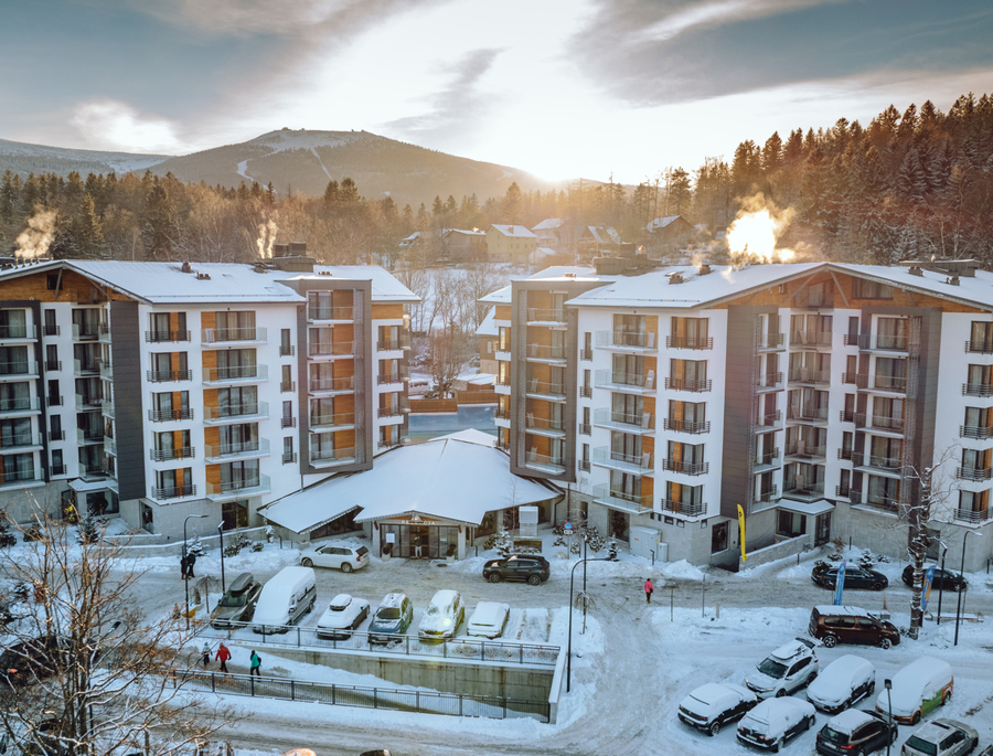 Moderní alpský hotelový komplex s balkony, zasněženým parkovištěm a kouřícími komíny, východ slunce nad zalesněnými horami a sjezdovkami
