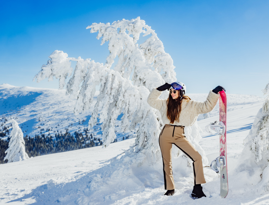 Smiling woman in helmet and winter gear poses with pink snowboard on sunny, snowy mountain slope by frosted trees