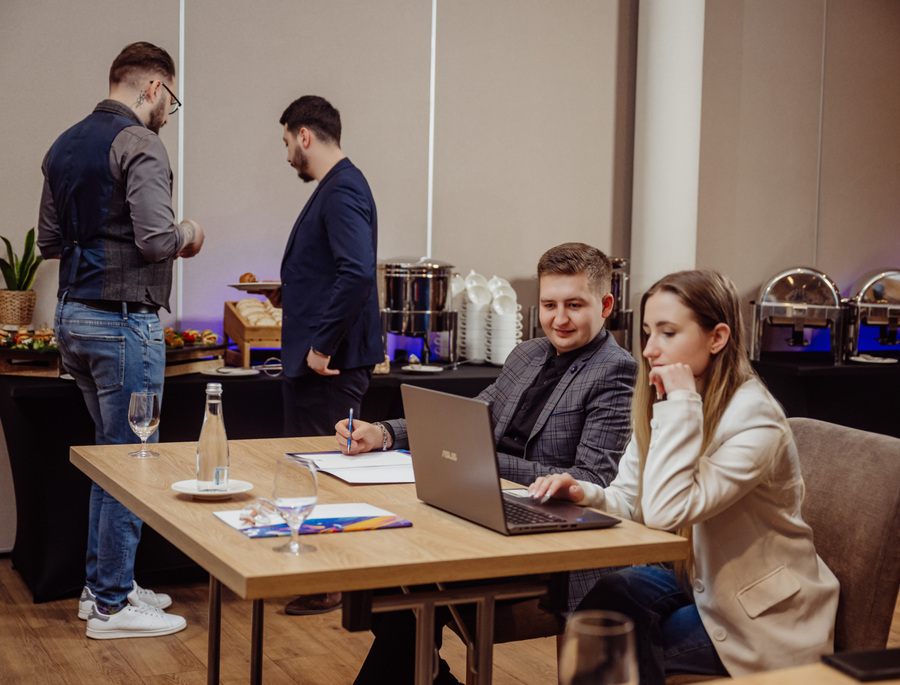 Konferenční místnost: dva kolegové u stolu s notebookem a poznámkami, raut s ohřívači a pečivem v pozadí.
