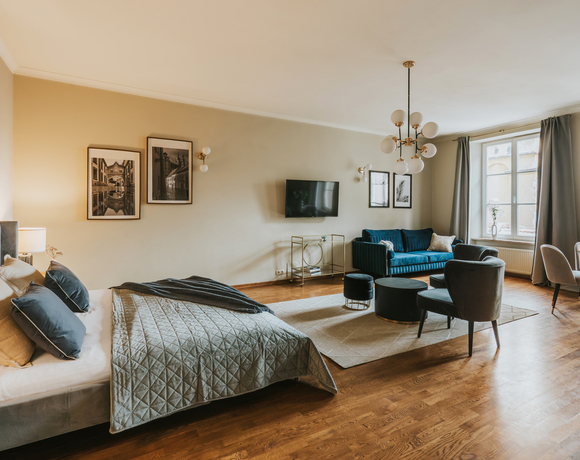Spacious studio suite with king bed, quilted bedding, teal velvet sofa, lounge chairs, wall TV, dining table and large bright window.