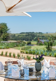 Umbrella-shaded terrace with glass dining table set with folded napkins, wine glasses and vase, overlooking rolling countryside and stone wall.