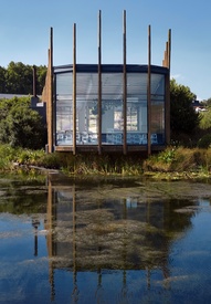 Glass-walled lakeside dining pavilion with wooden poles, indoor seating and reflections on a tranquil pond and gardens.