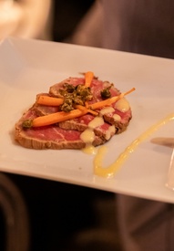 Server presenting seared beef carpaccio with baby carrots, capers and mustard drizzle on a square plate in candlelit dining