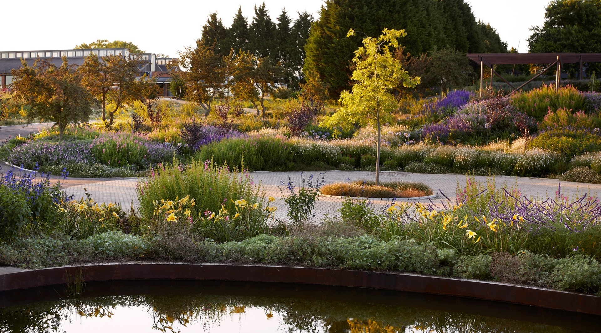 Sunlit hotel gardens with wildflower beds, circular paved driveway, young trees and reflective pond creating tranquil grounds.