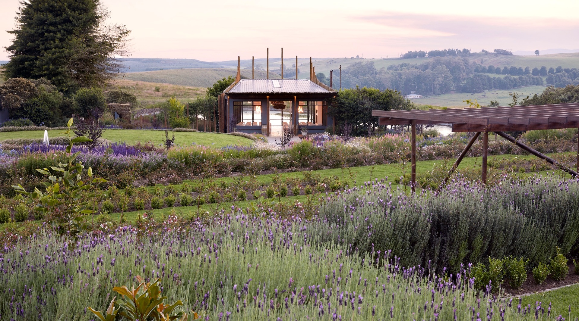 Glass-front spa pavilion amid lavender gardens, pergola and fountain, overlooking a pond and rolling countryside.