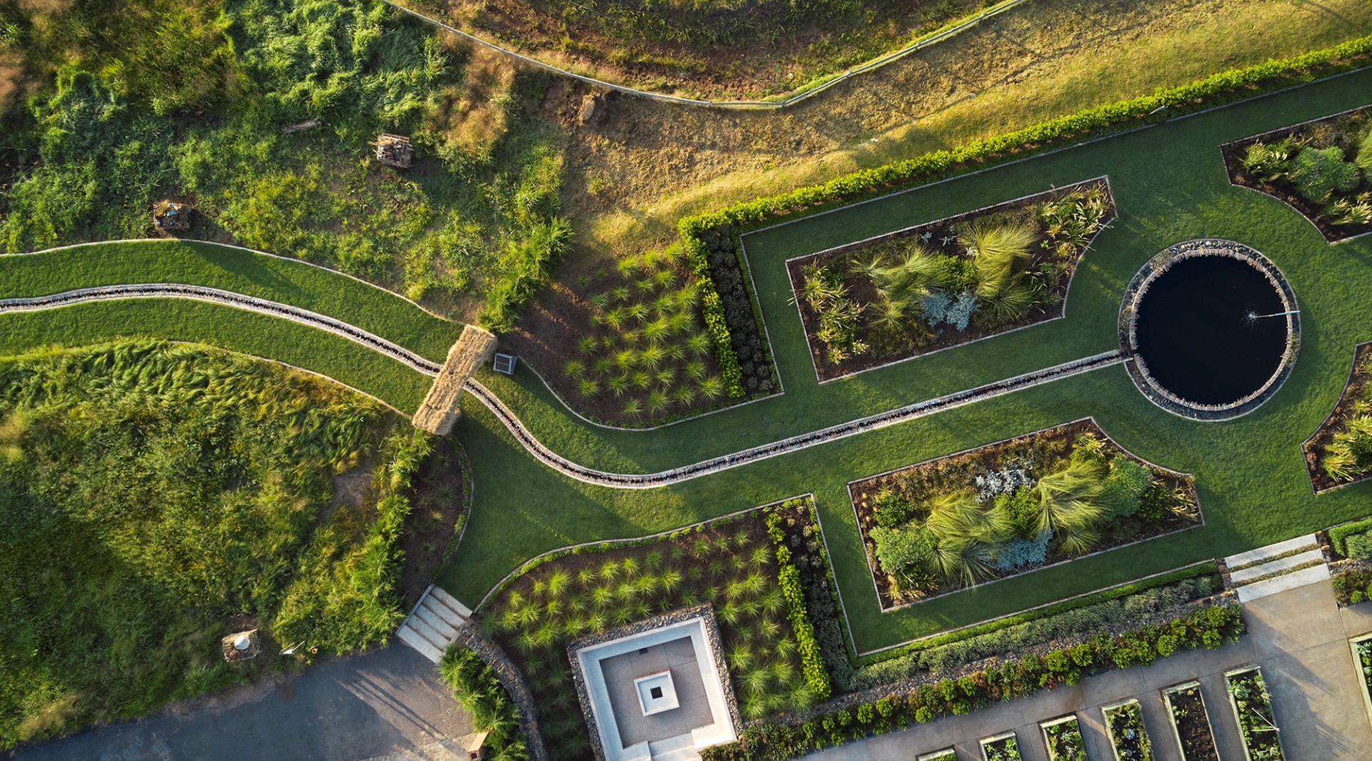 Aerial hotel gardens with geometric lawns, planted beds, winding paths and a circular reflecting pond with a small fountain.