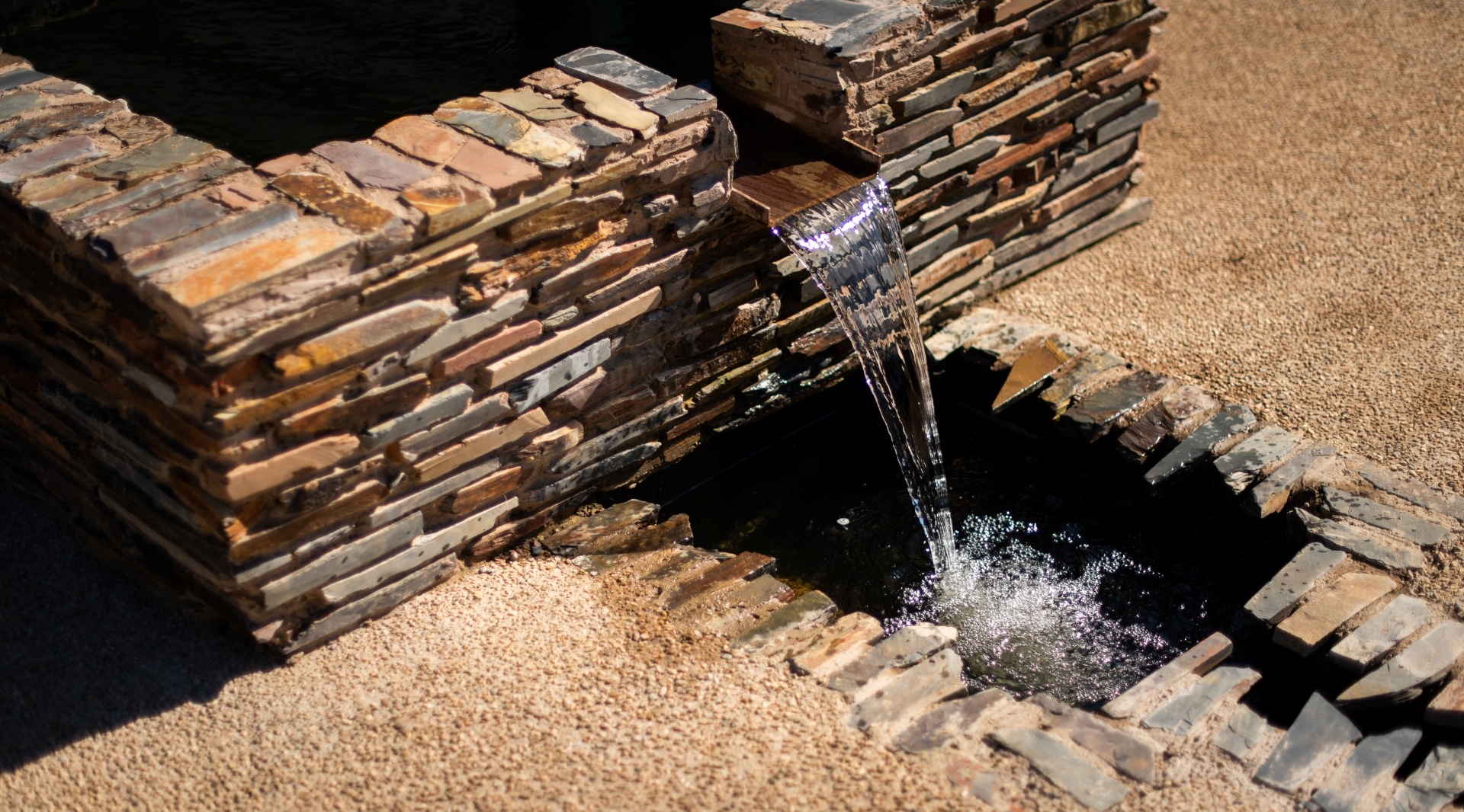 Sunlit stacked-stone water feature with a narrow cascade flowing into a dark basin, outdoor courtyard detail.